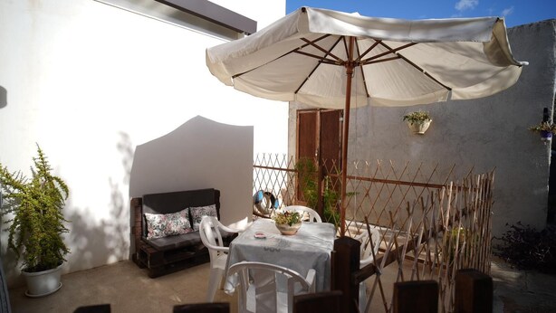 A patio with a table and chairs, a sofa, a large umbrella, and potted plants. - The House of Flowers, in the heart of the Gulf of Macari 2.5 km from the sea. (Castelluzzo)