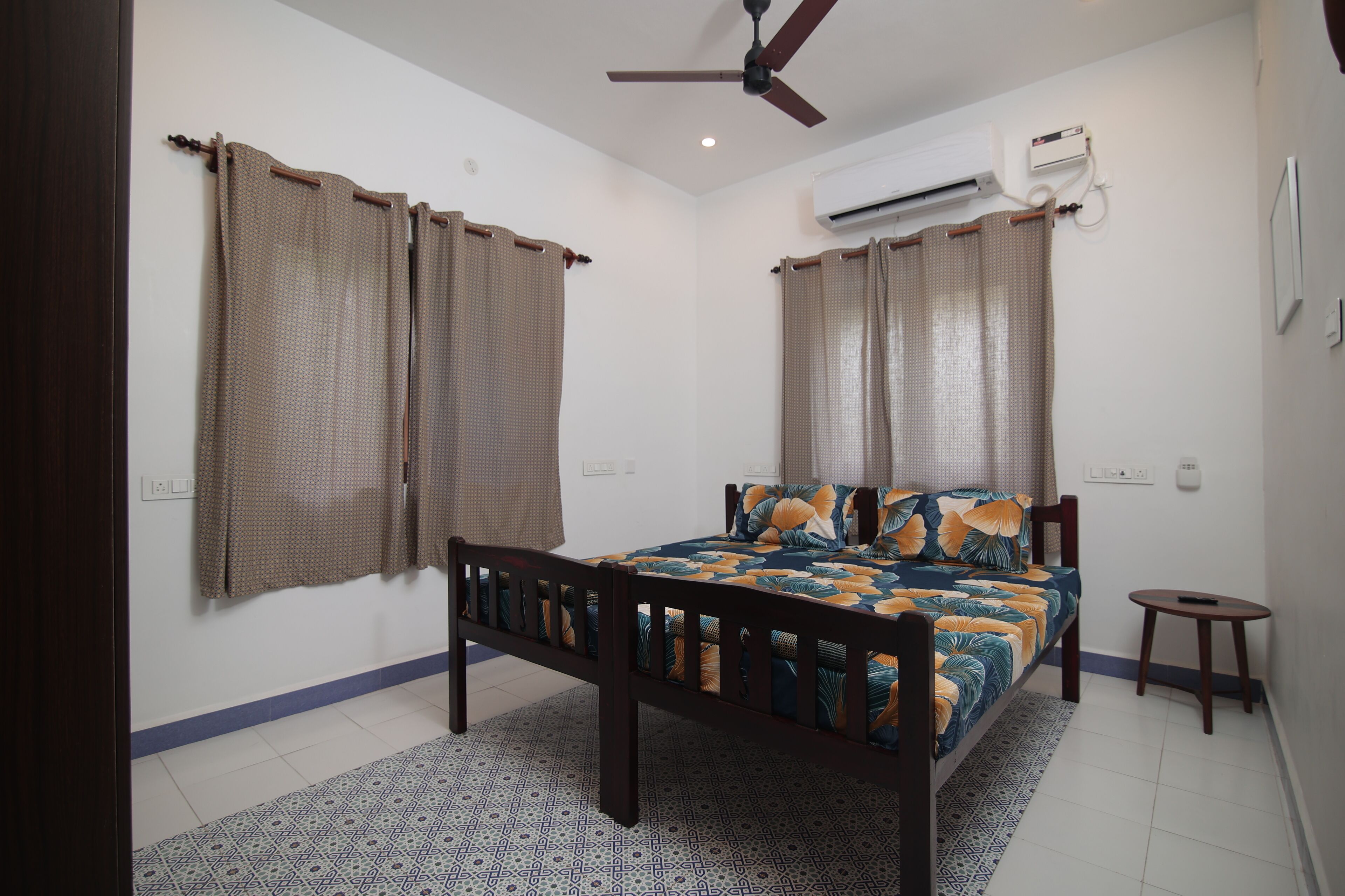 A bedroom with a bed, curtains, a ceiling fan, an air conditioning unit, and a small table.