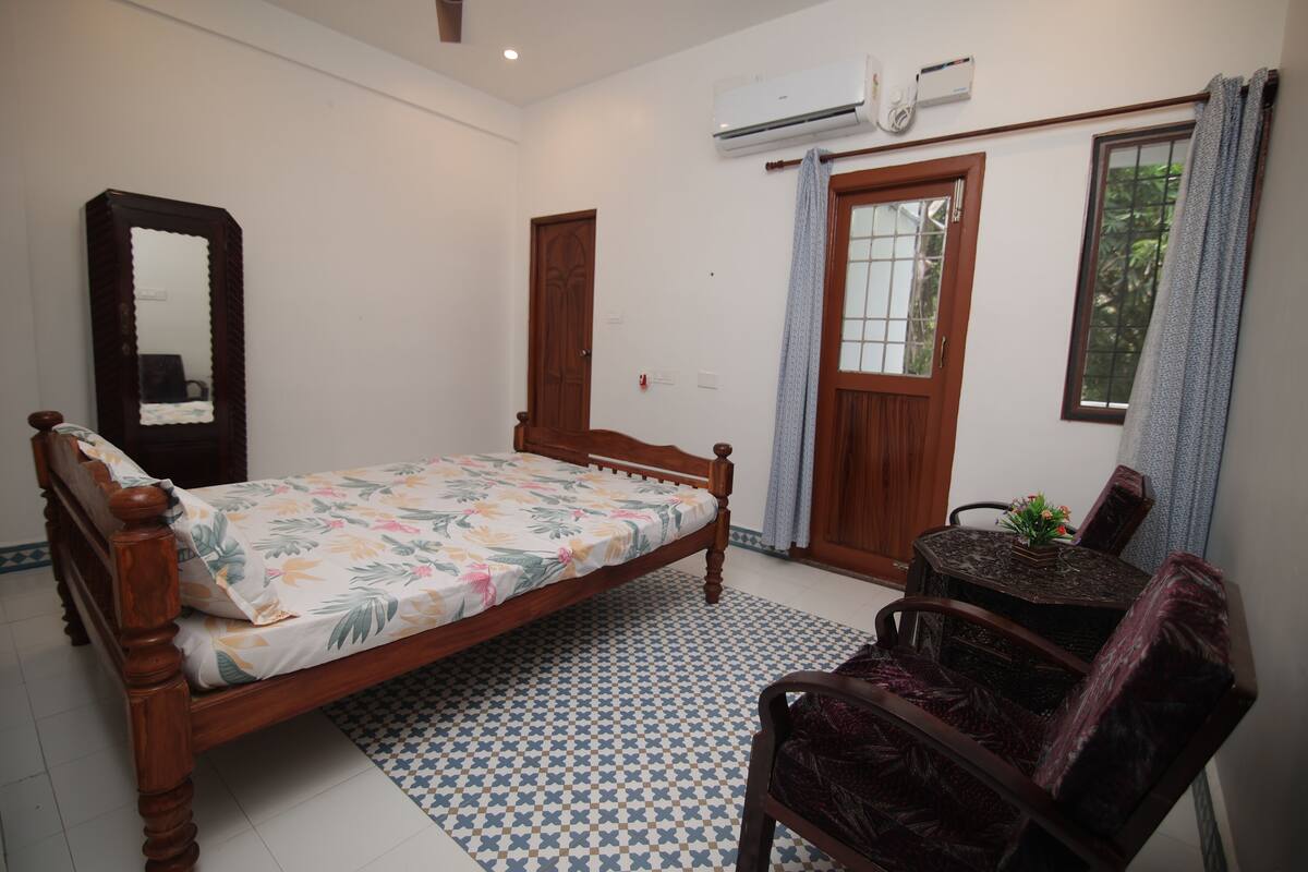 A bedroom with a wooden bed, a small round table with a plant, a chair, a mirror, and a window with curtains.