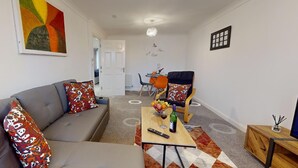 Una sala moderna con un sofá gris, cojines de colores, una mesa de centro de madera con un control remoto y una botella, y un televisor sobre un mueble de madera.