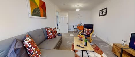 A modern living room with a grey sofa, colorful cushions, a wooden coffee table with a remote control and a bottle, and a TV on a wooden stand.