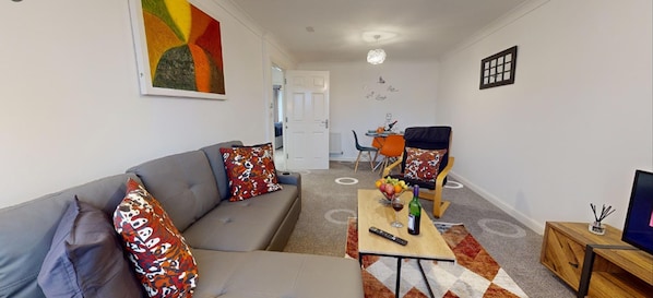 A modern living room with a grey sofa, colorful cushions, a wooden coffee table with a remote control and a bottle, and a TV on a wooden stand.