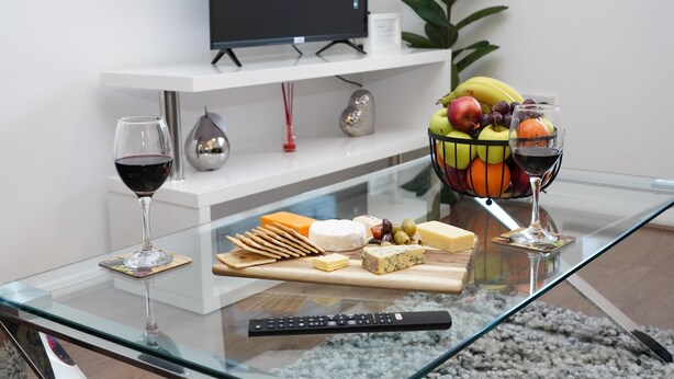 A glass table with a cheese board, fruits, and wine glasses. A remote control is on the table. - 4guests-parking-2br-familyfriendly (London)