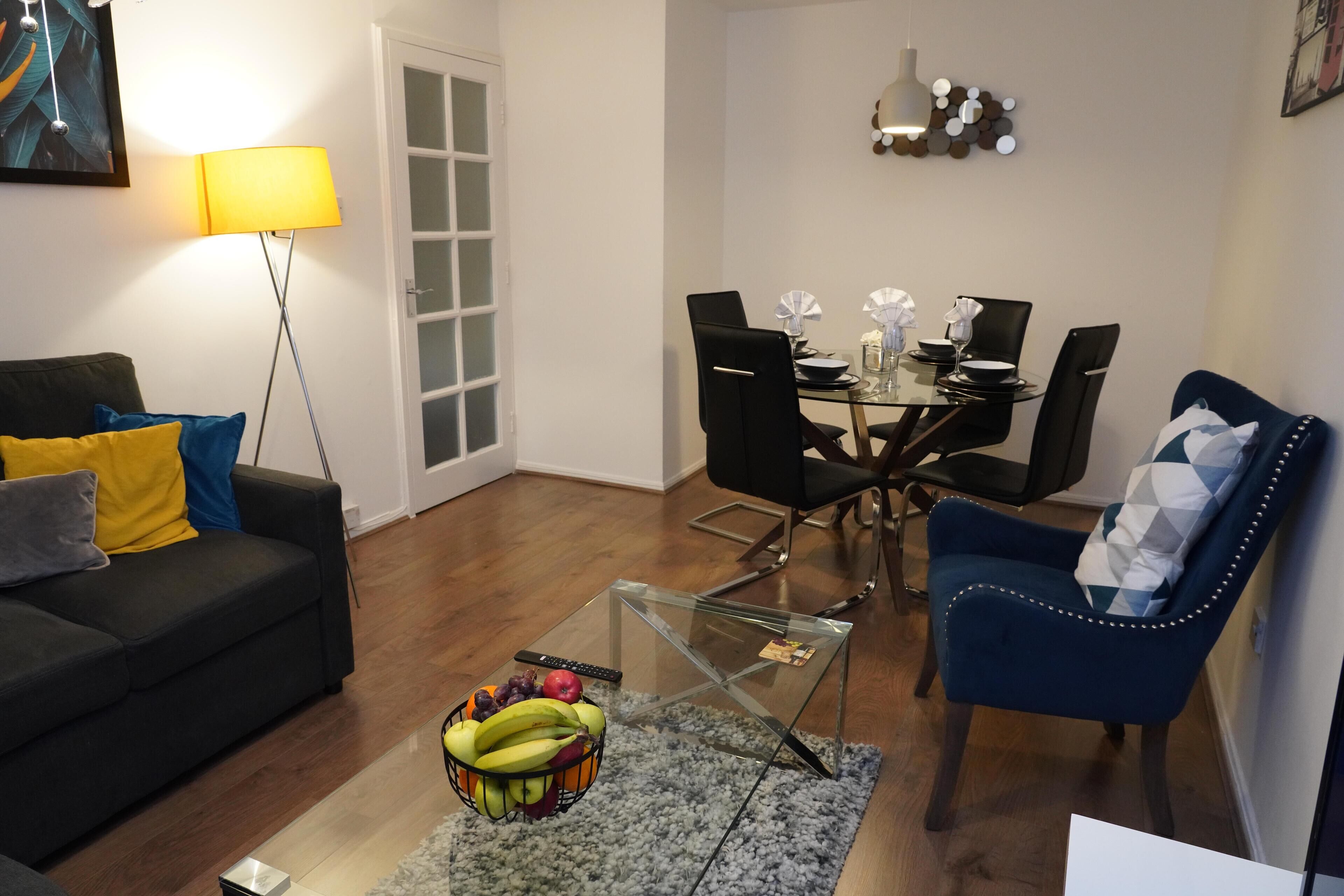 A living room with a sofa, a glass coffee table with fruit, a dining area with chairs, and a lamp.