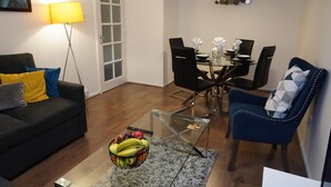 A living room with a sofa, a glass coffee table with fruit, a dining area with chairs, and a lamp. - 4guests-parking-2br-familyfriendly (London)
