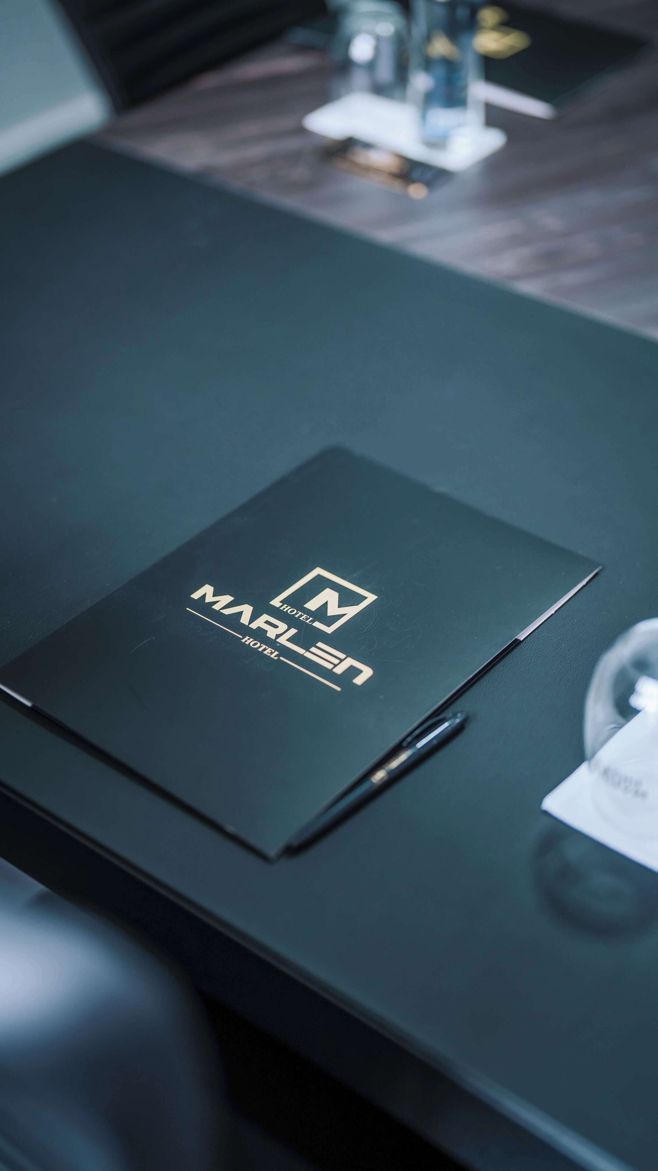 Una cartella nera con il logo dell'Hotel Marlen su una scrivania in legno scuro.