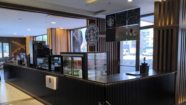 A Starbucks coffee shop with a modern design, including a counter with a glass display case, a menu board, and a sign indicating the direction to the coffee area. - Marlen Hotel Denizli (Denizli)