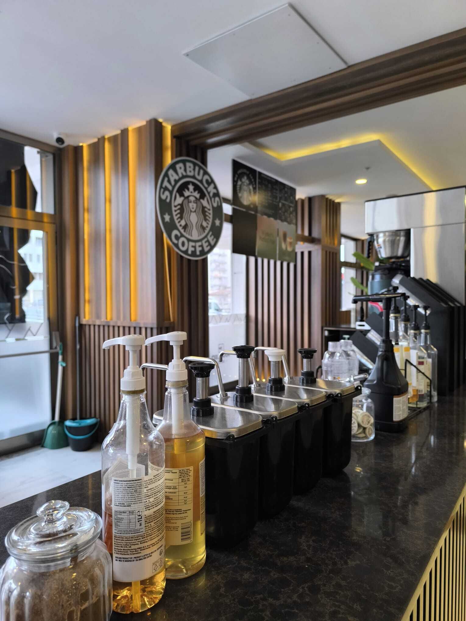 Interior de uma cafeteria Starbucks, com um balcão exibindo álcool em gel, uma máquina de café e diversas garrafas.