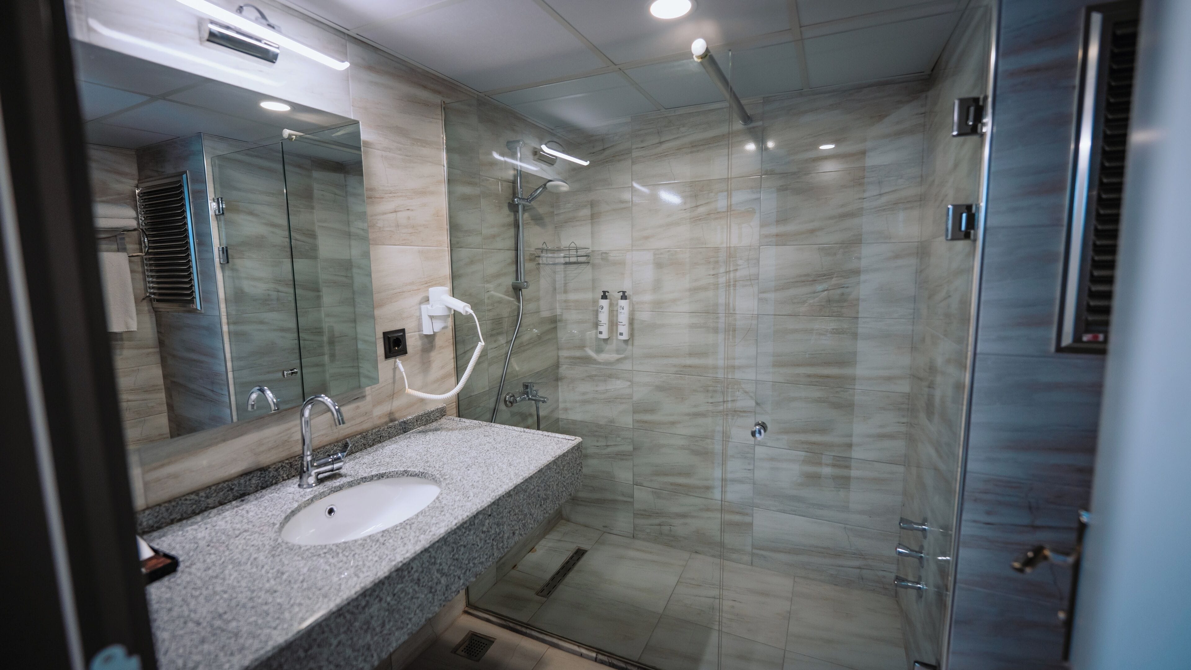 A modern bathroom with a granite countertop, a glass shower enclosure, and a large mirror.
