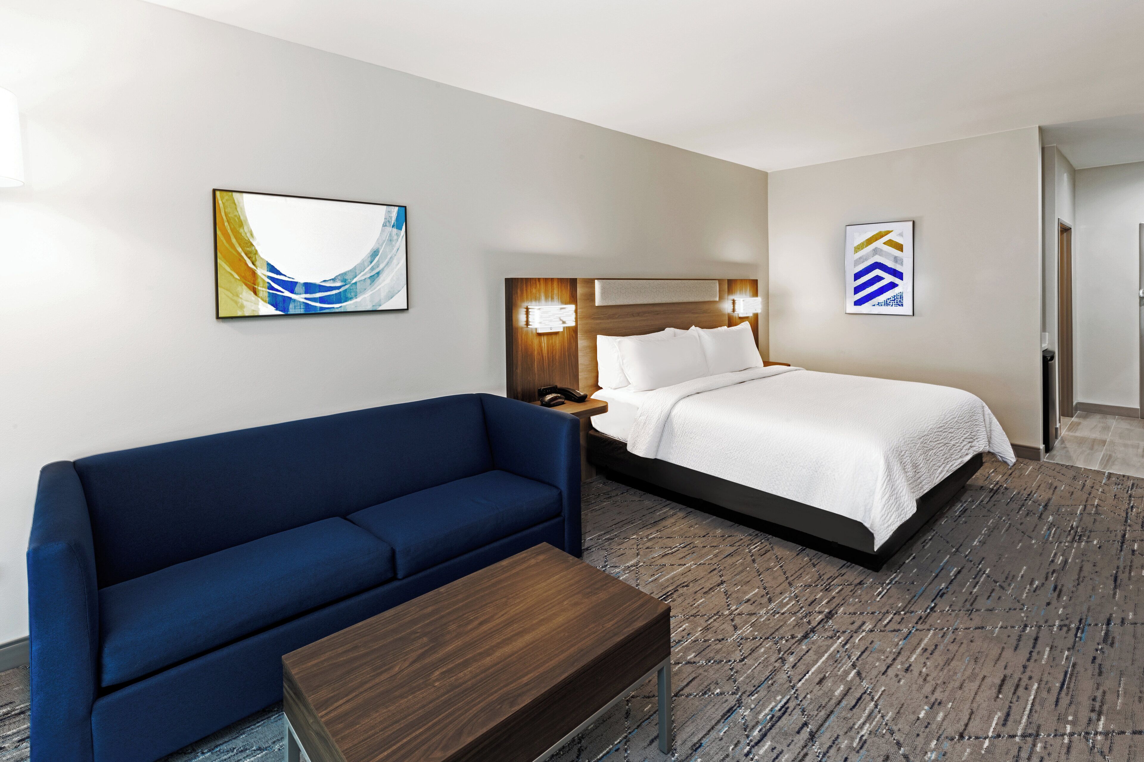 A hotel room with a blue sofa, a wooden coffee table, a bed with white bedding, and abstract wall art.