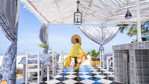 Una terrazza fronte spiaggia con pavimento a scacchi, tavoli bianchi con tovaglioli a motivi blu e bianchi, e una persona che indossa un abito giallo.