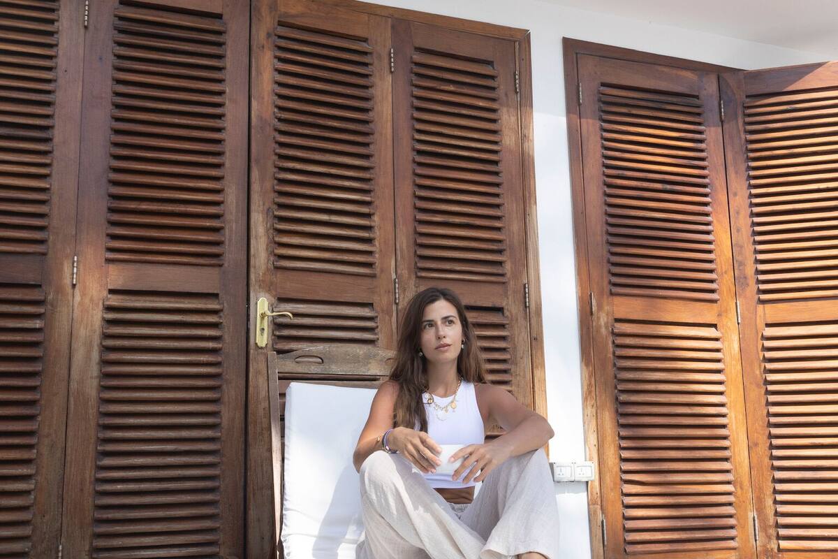 Una persona sentada en un banco de madera con un colchón blanco, frente a contraventanas de madera, y una palmera al fondo.