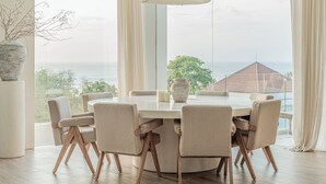 A modern dining room with a round table, six chairs, a hanging light fixture, and a large window offering a view of the sea and buildings. - Villa Namiko by Alfred in Bali (Pecatu)