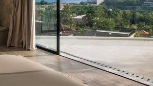 A modern hotel room with a large sliding glass door leading to a balcony with a view of the sea and a distant building. - Villa Namiko by Alfred in Bali (Pecatu)
