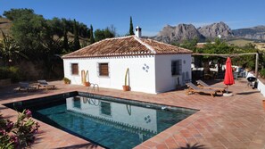 Zona junto a la piscina con un edificio blanco, una alberca y montañas al fondo.