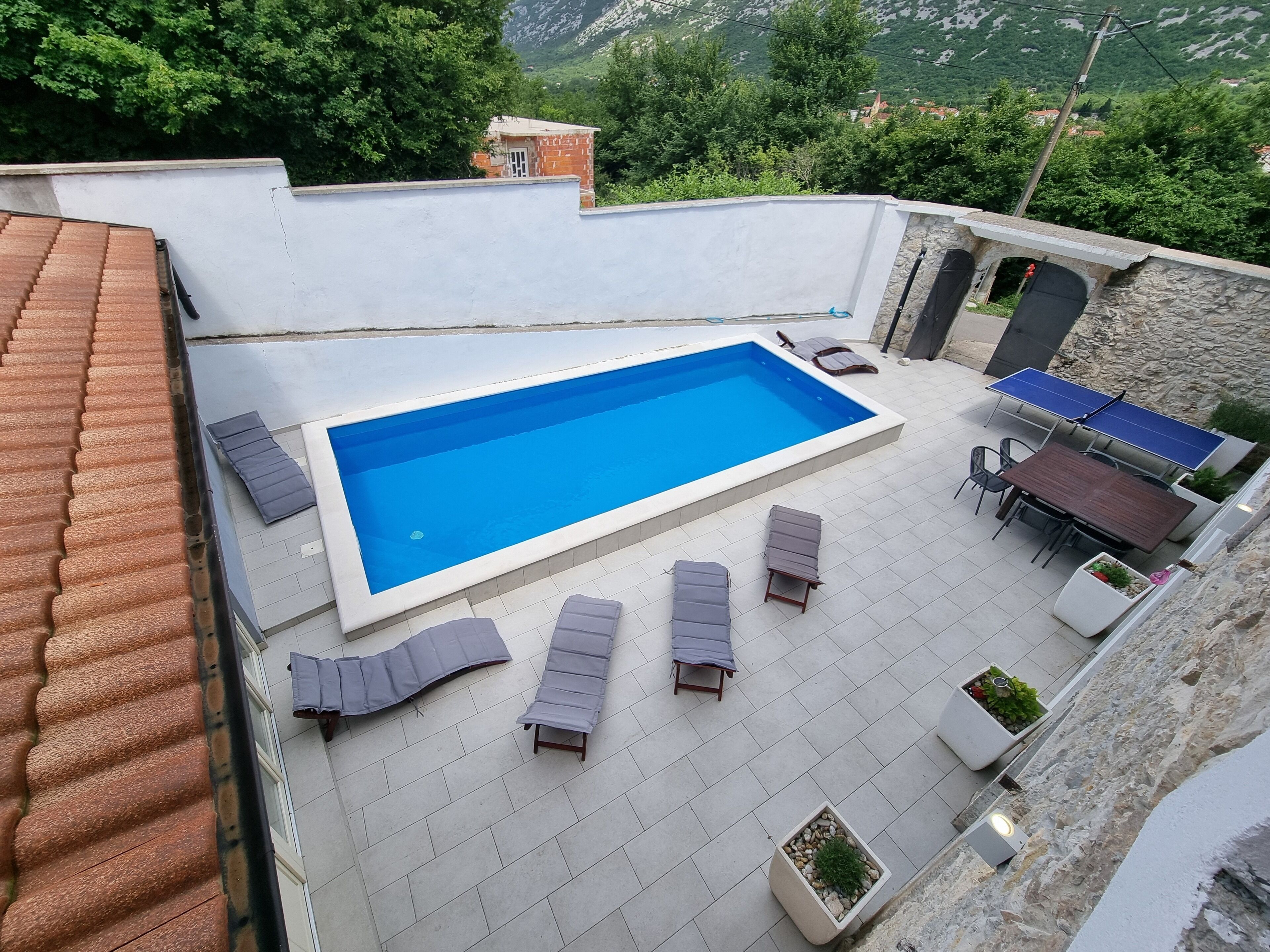Una piscina en la azotea con sillas de descanso y una mesa, rodeada de plantas en macetas y un muro de piedra.