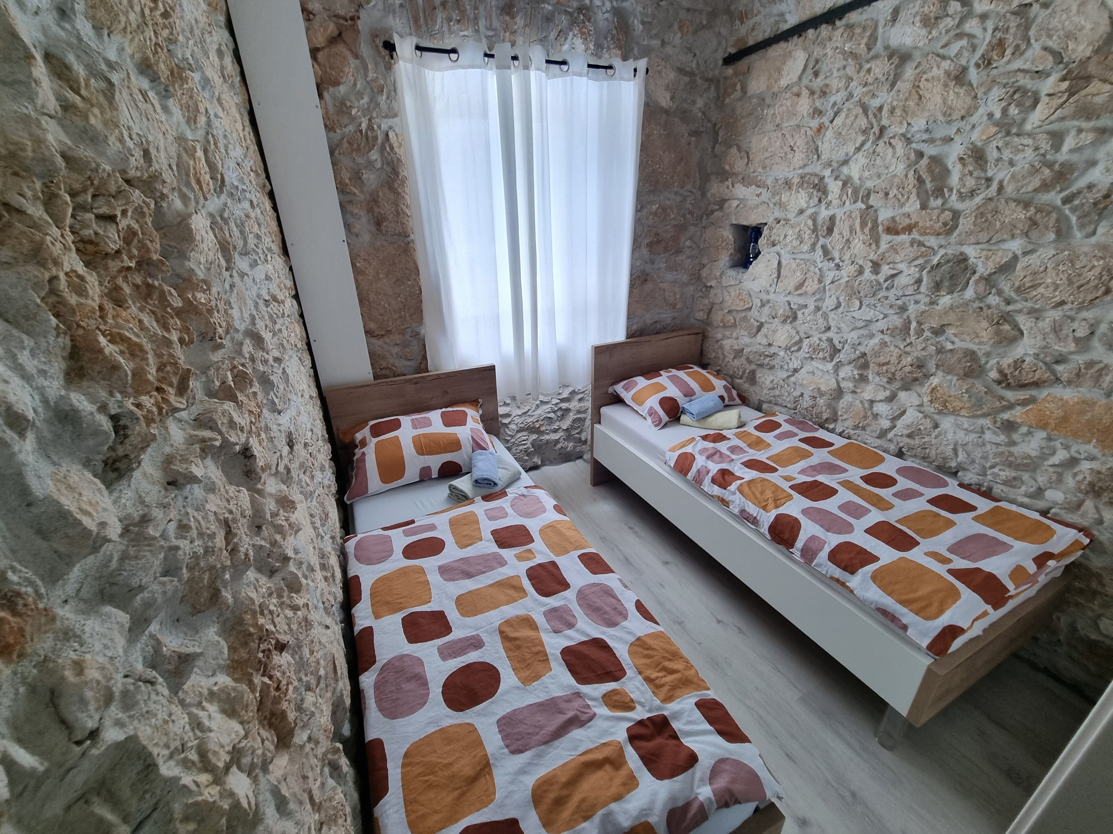 A small, rustic room with two wooden beds, stone walls, and a window with white curtains.