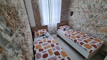 A small, rustic room with two wooden beds, stone walls, and a window with white curtains.
