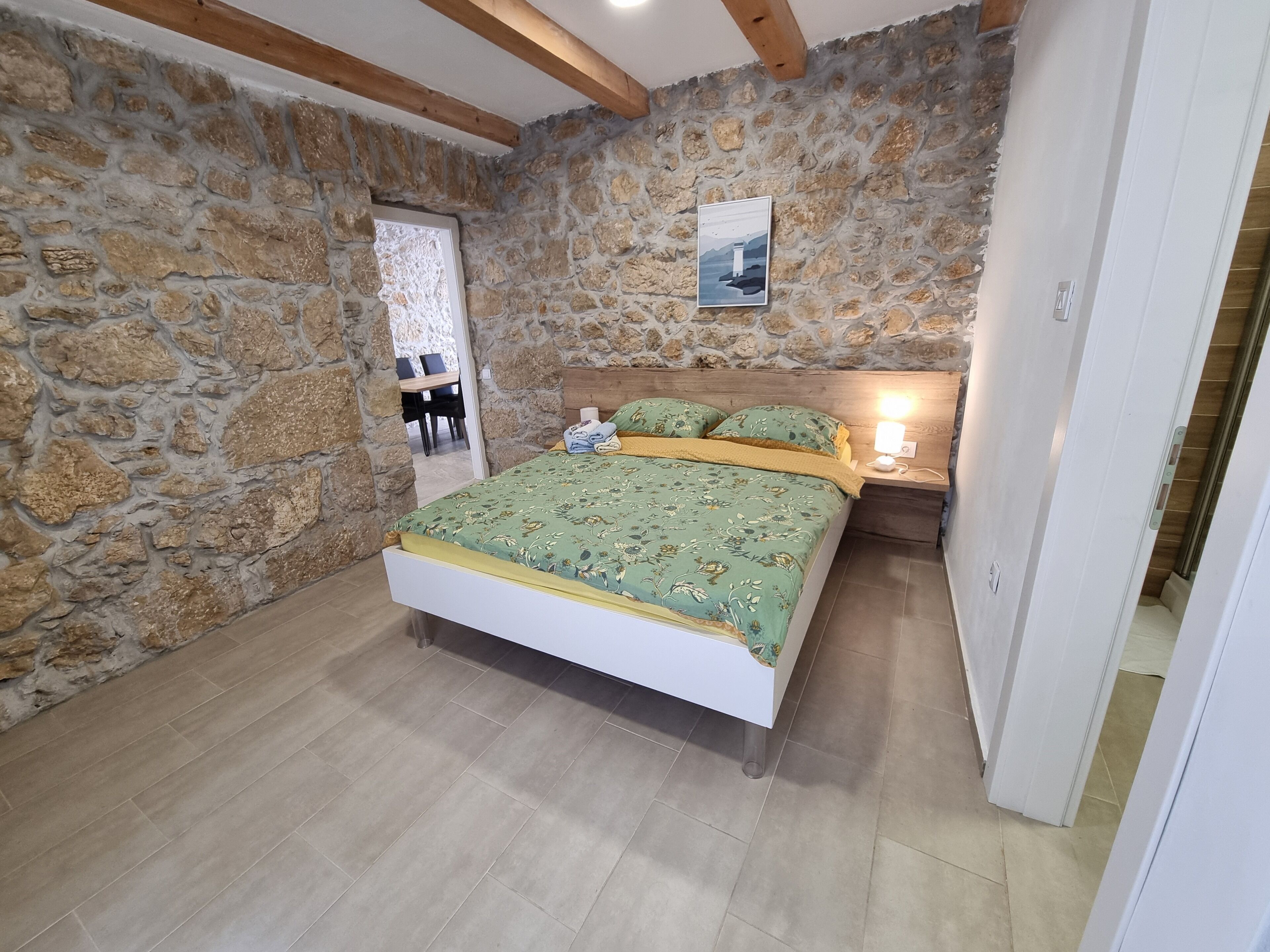 A bedroom with a stone wall, a wooden ceiling, a bed with floral bedding, a nightstand with a lamp, and a door leading to another room.