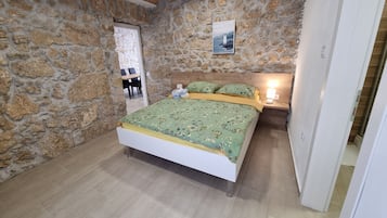 A bedroom with a stone wall, a wooden ceiling, a bed with floral bedding, a nightstand with a lamp, and a door leading to another room.