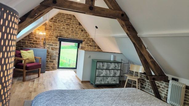 Une chambre mansardée chaleureuse, avec un plafond en pente, un mur en pierre, un lit avec une literie à motifs, un fauteuil avec des coussins, un radiateur et une vue sur l’extérieur par une fenêtre.