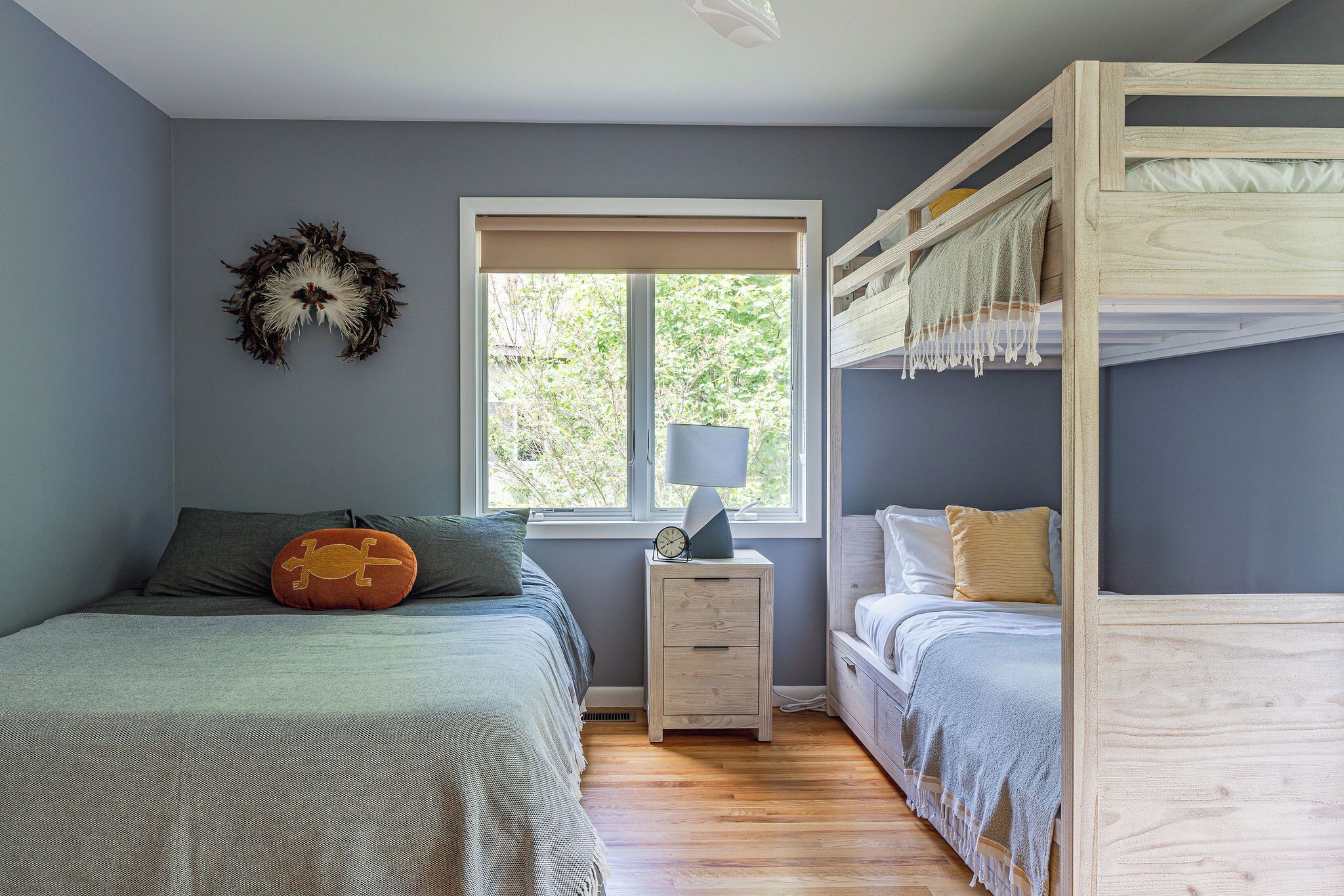 A bedroom with a bunk bed, a bedside table with a lamp, and a window with a view of trees.