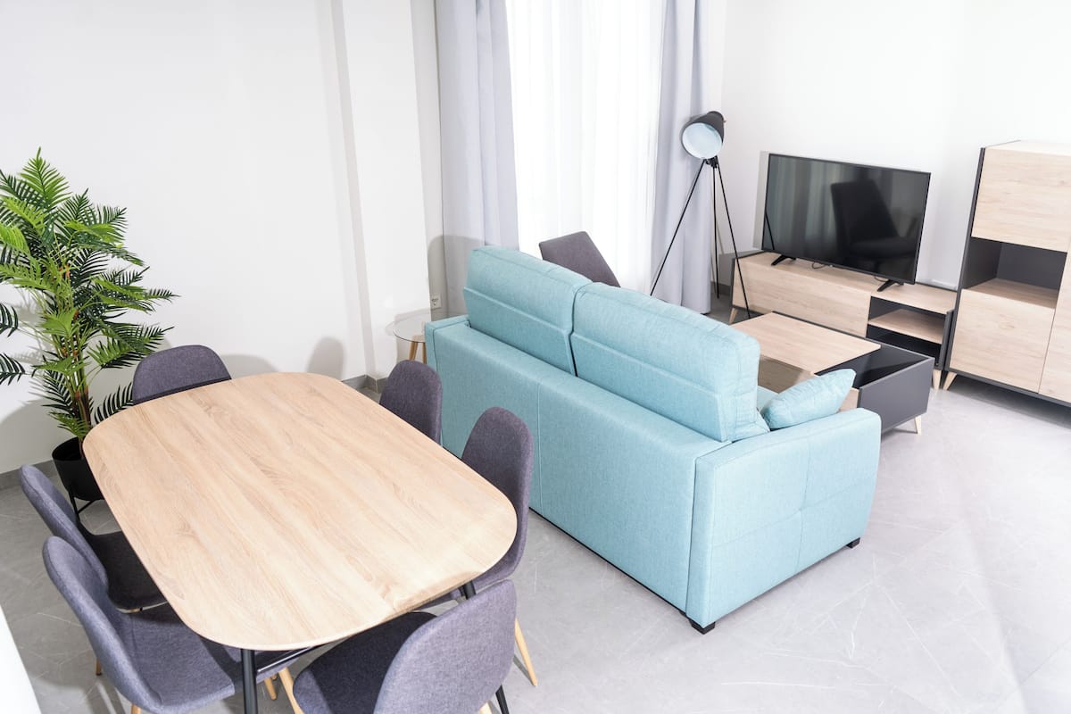 A modern living room with a light blue sofa, a wooden dining table, and a TV.