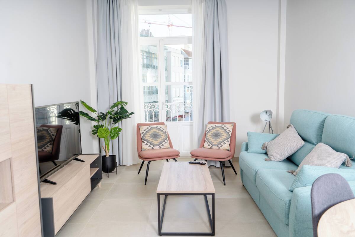 A modern living room with a sofa, two armchairs, a coffee table, a TV, and a large window with curtains.