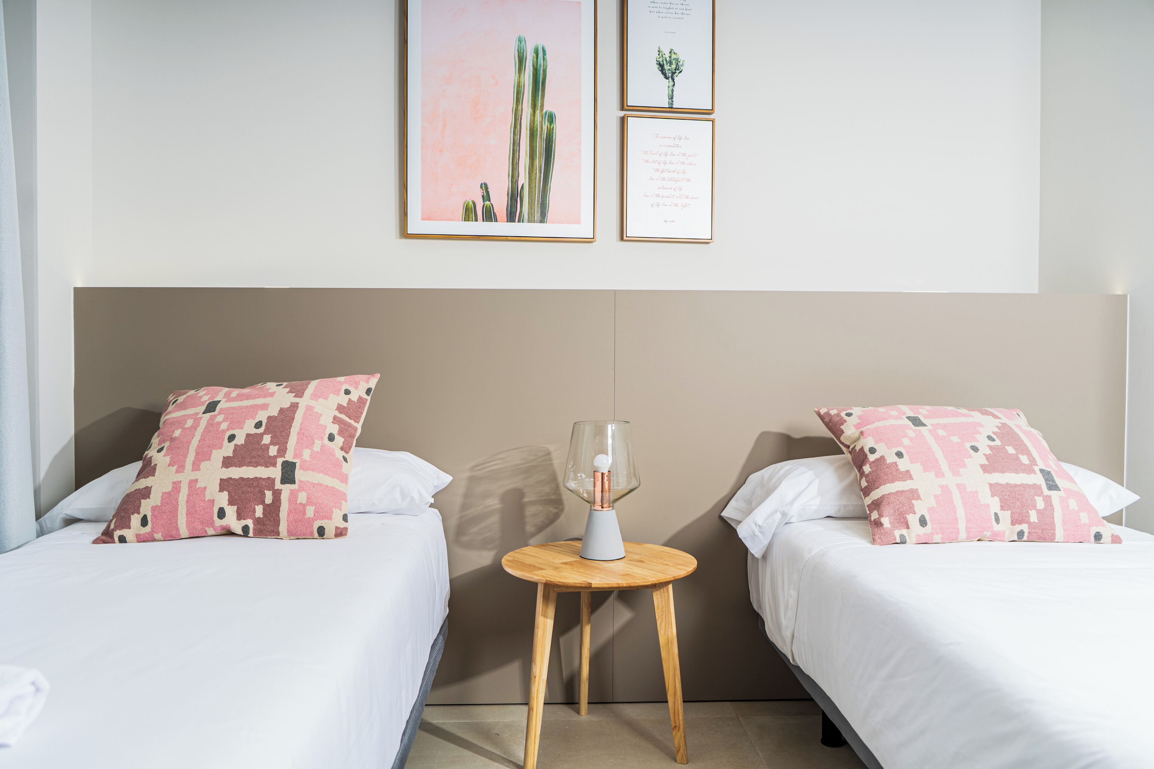 Una habitación de hotel con dos camas, cada una con un cojín estampado, una mesita de madera con un jarrón y cuadros en la pared.