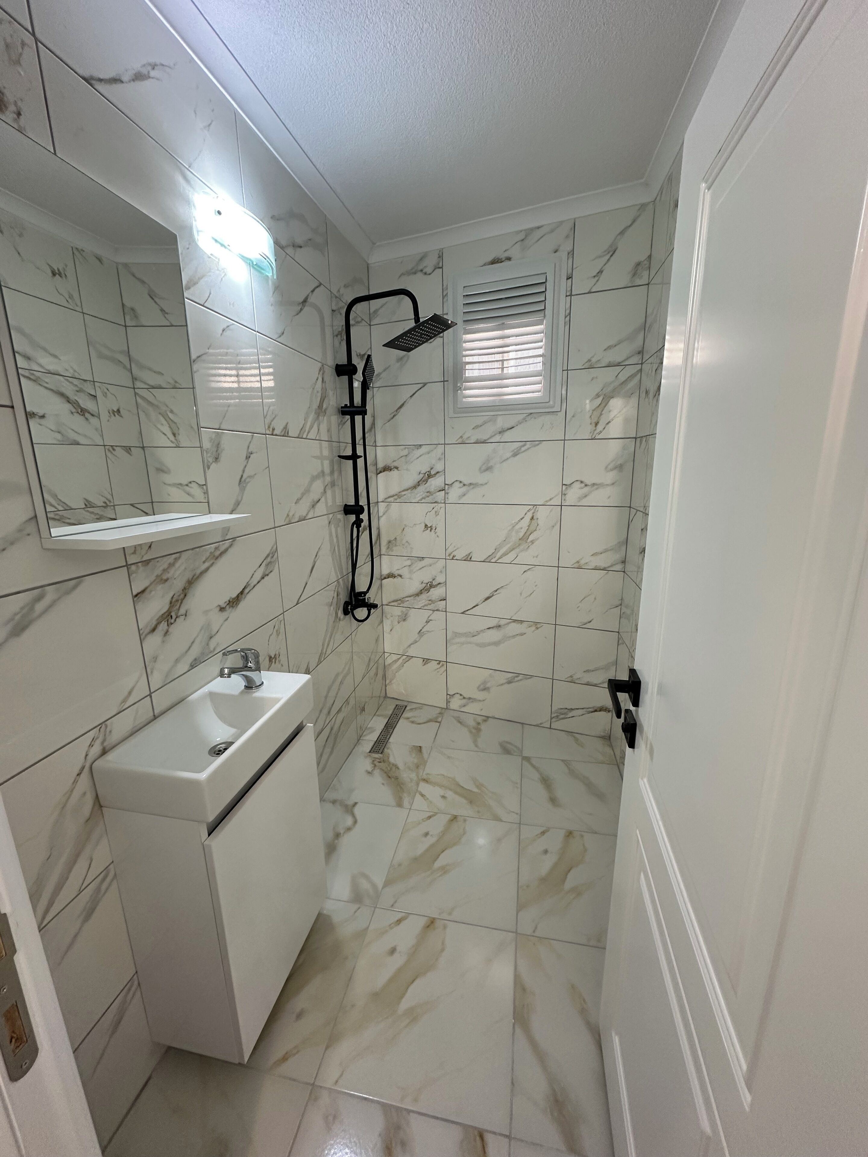 A bathroom with a white sink, a mirror, a shower with a handheld showerhead, and a window with blinds.