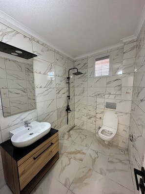 A bathroom with a marble-tiled floor and walls, a white sink with a wooden vanity, a handheld showerhead, and a toilet.