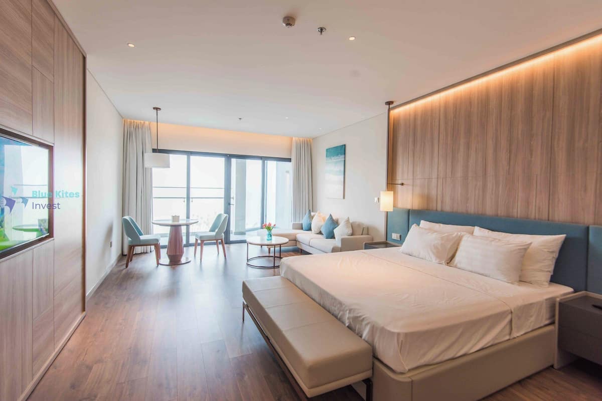 Une chambre d’hôtel moderne dotée d’un grand lit, d’un coin salon avec un canapé et des fauteuils, d’une petite table et d’un téléviseur fixé au mur.