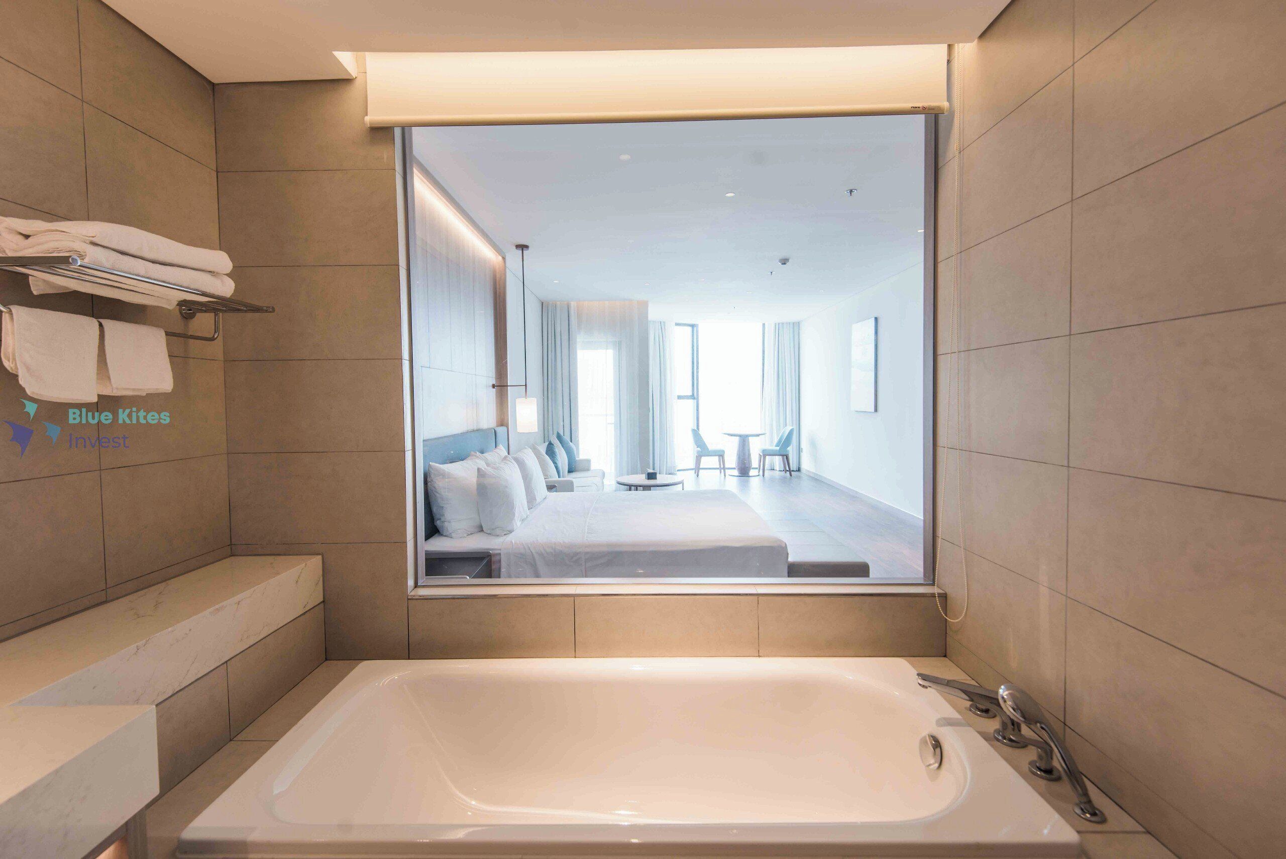 A modern bathroom with a large bathtub, a towel rack, and a mirror reflecting a bedroom with a bed and a seating area.