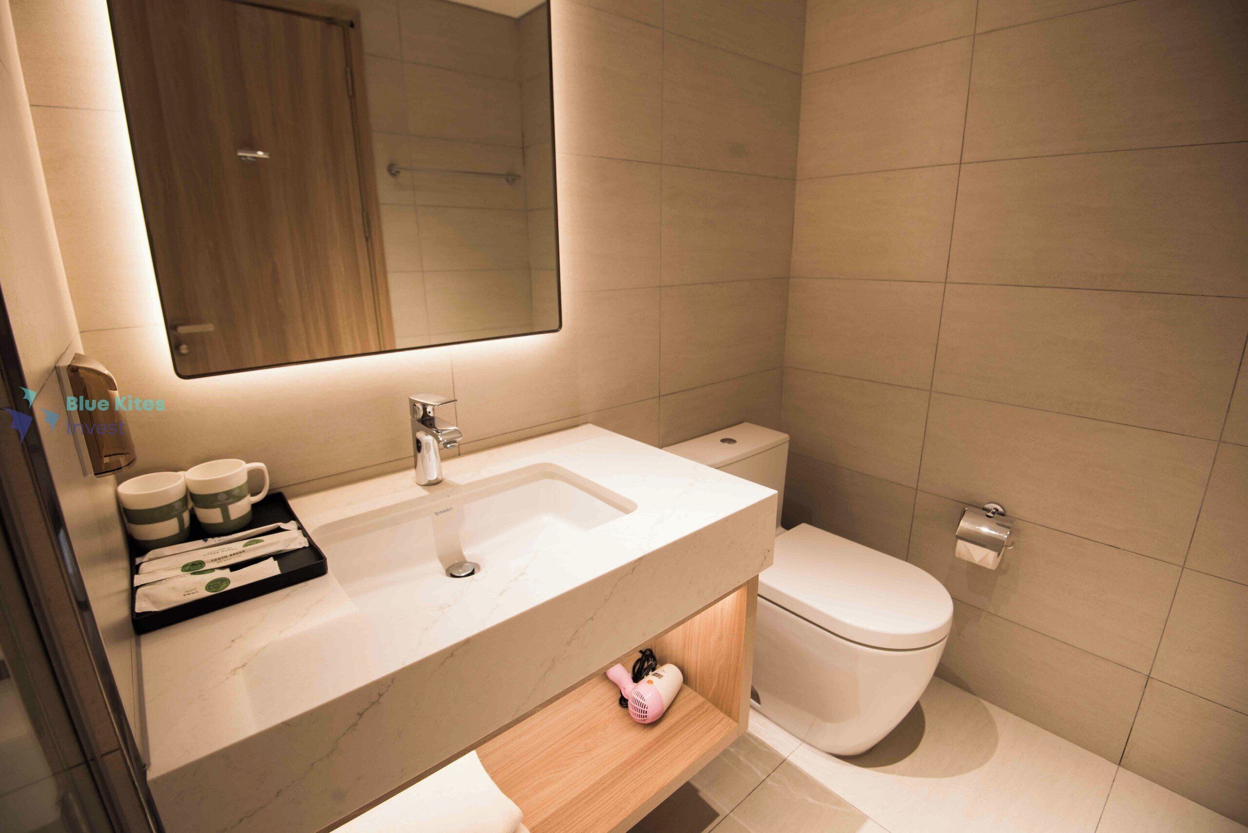 A modern bathroom with a white sink, mirror, and toilet. There is a tray with toiletries and a hairdryer on the sink.