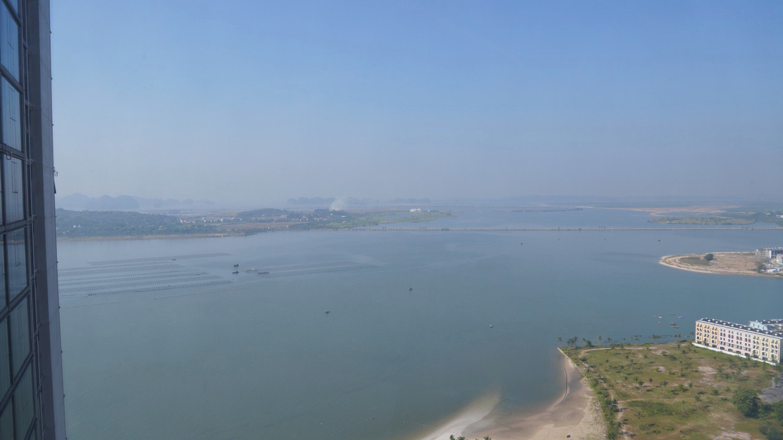 Vista costera con una extensa masa de agua, una playa arenosa y un edificio de varios pisos.