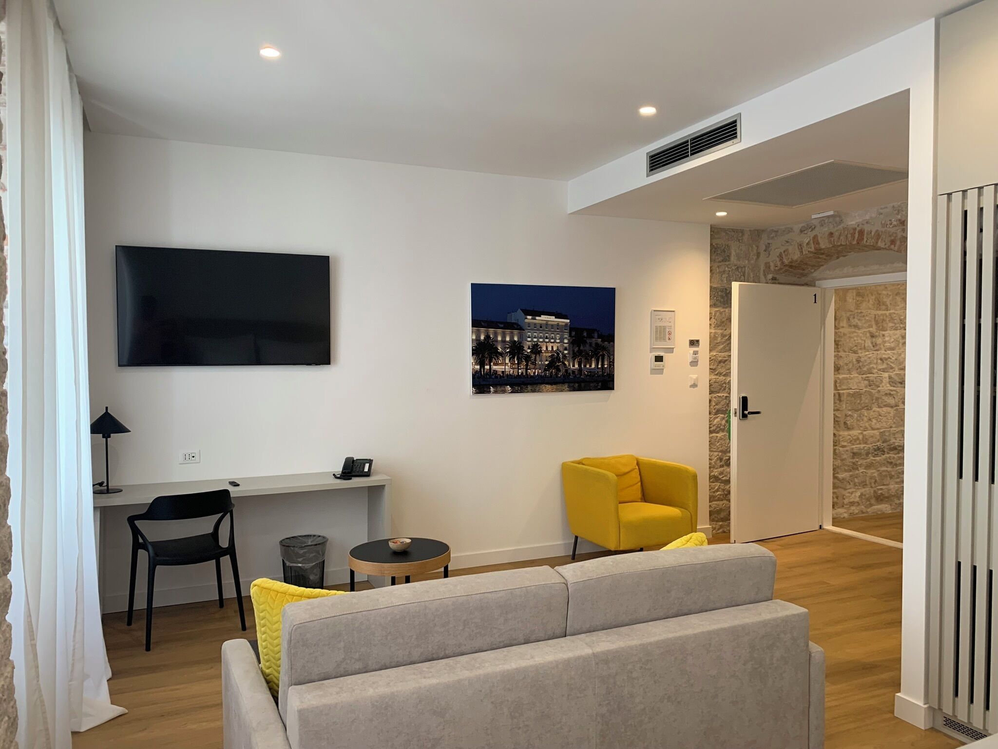 A modern living room with a grey sofa, a yellow armchair, a flat-screen TV, a desk with a phone, and a painting of a cityscape.