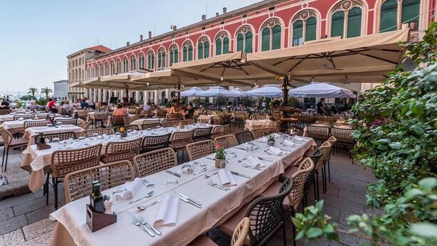 An outdoor dining area with tables and chairs, a red building with arched windows, and umbrellas providing shade. - Priuli Heritage Suites (Split)