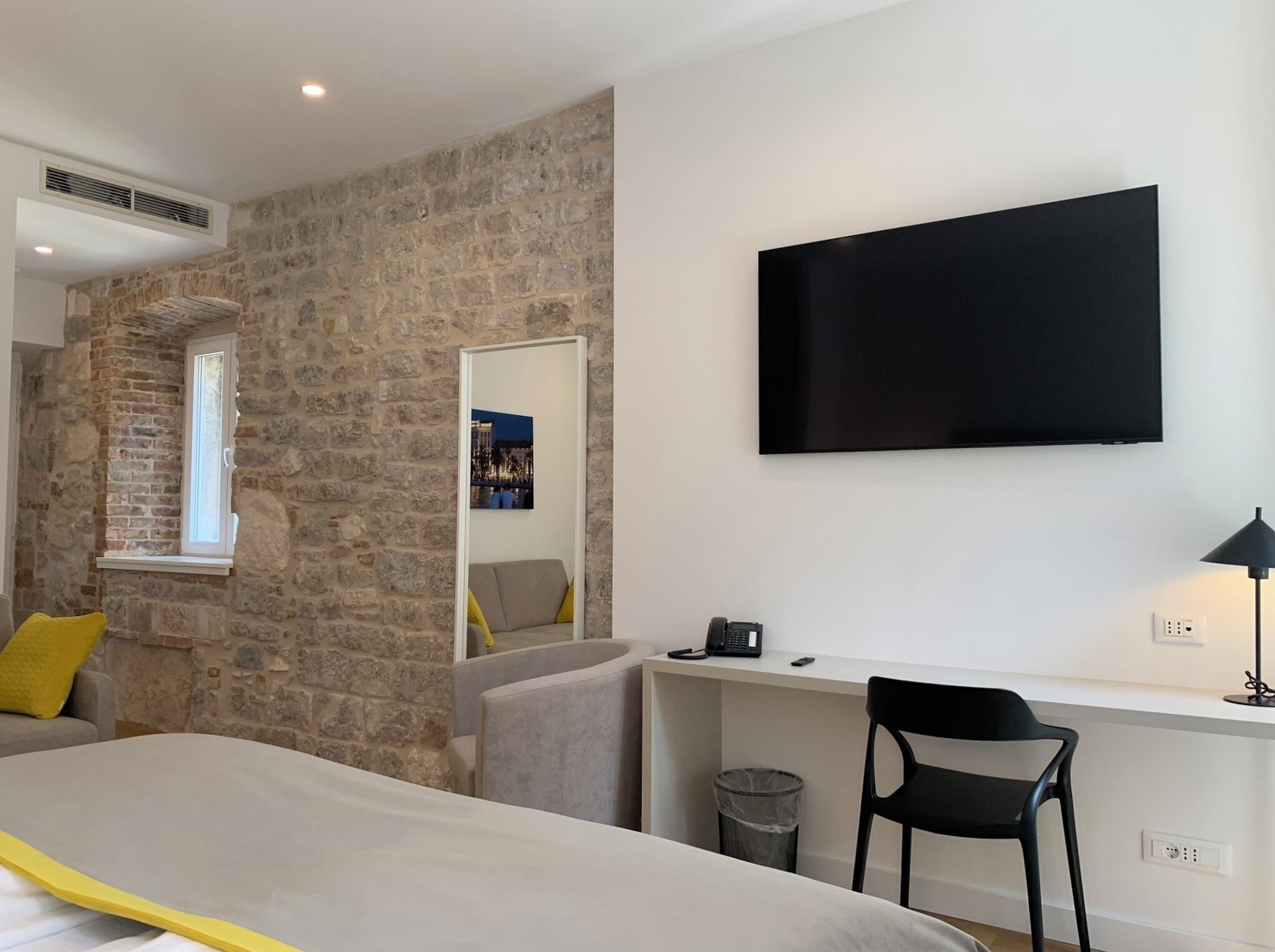 A modern hotel room with a stone wall, a bed with a yellow accent, a desk with a chair, a TV, and a trash bin.