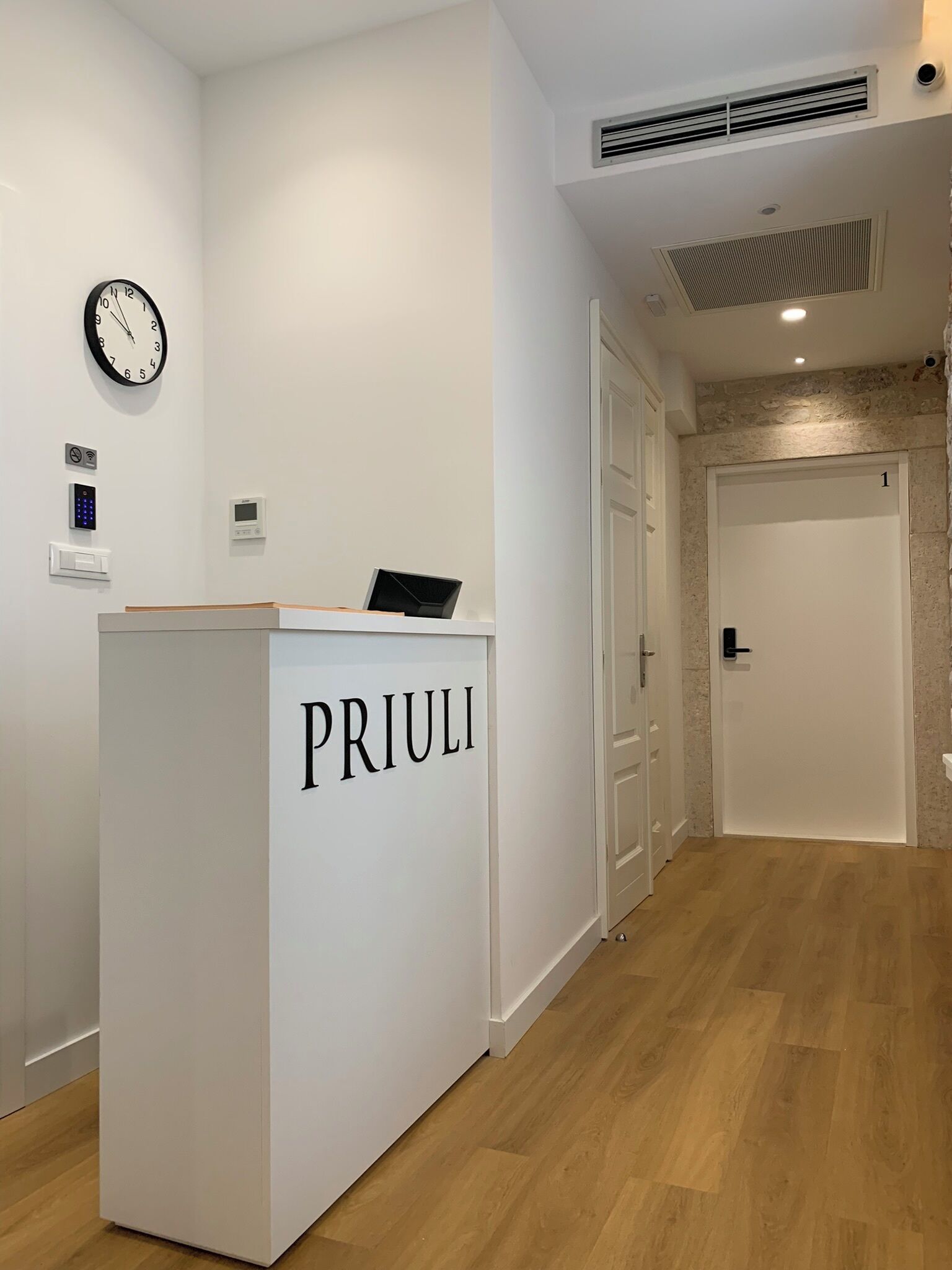 A reception desk labeled "PRIULI" with a clock, a digital control panel, and a door with a number "1".