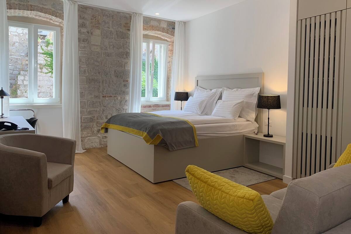 Ein modernes Schlafzimmer mit Steinwand, einem Bett mit weißer Bettwäsche, zwei Nachttischlampen und Blick durch ein Fenster.