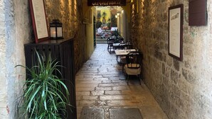 A narrow stone corridor with tables and chairs, potted plants, and framed menus on the walls. - Priuli Heritage Suites (Split)