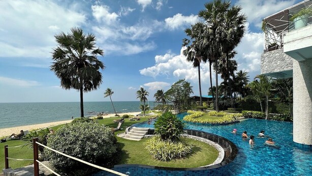 Ein Pool im Resort mit Blick auf Strand und Meer, umgeben von Palmen und einem Steinhaus.