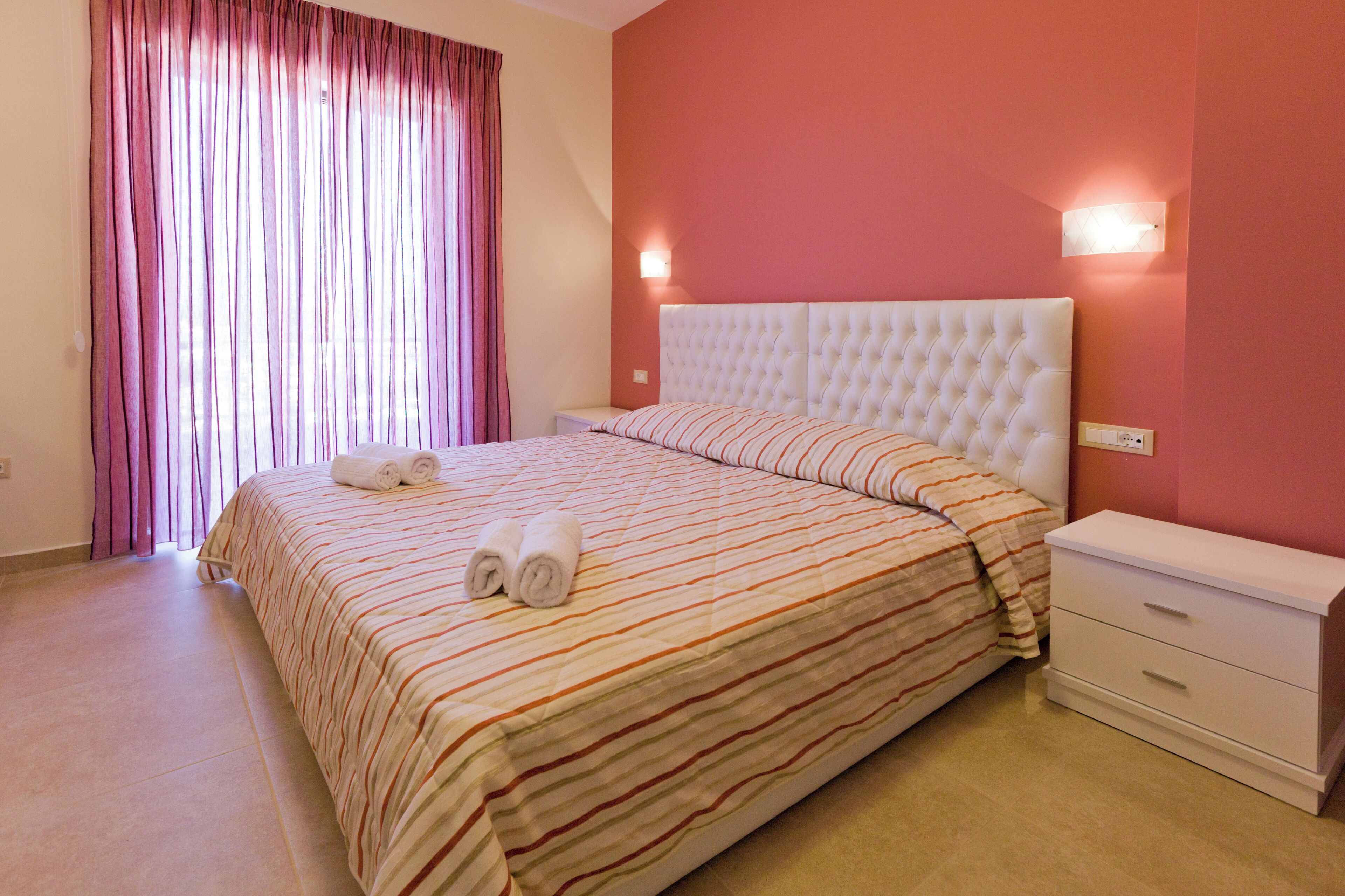Un dormitorio con una cama grande, ropa de cama a rayas, un cabecero blanco, una mesita de noche y una lámpara fijada a la pared.
