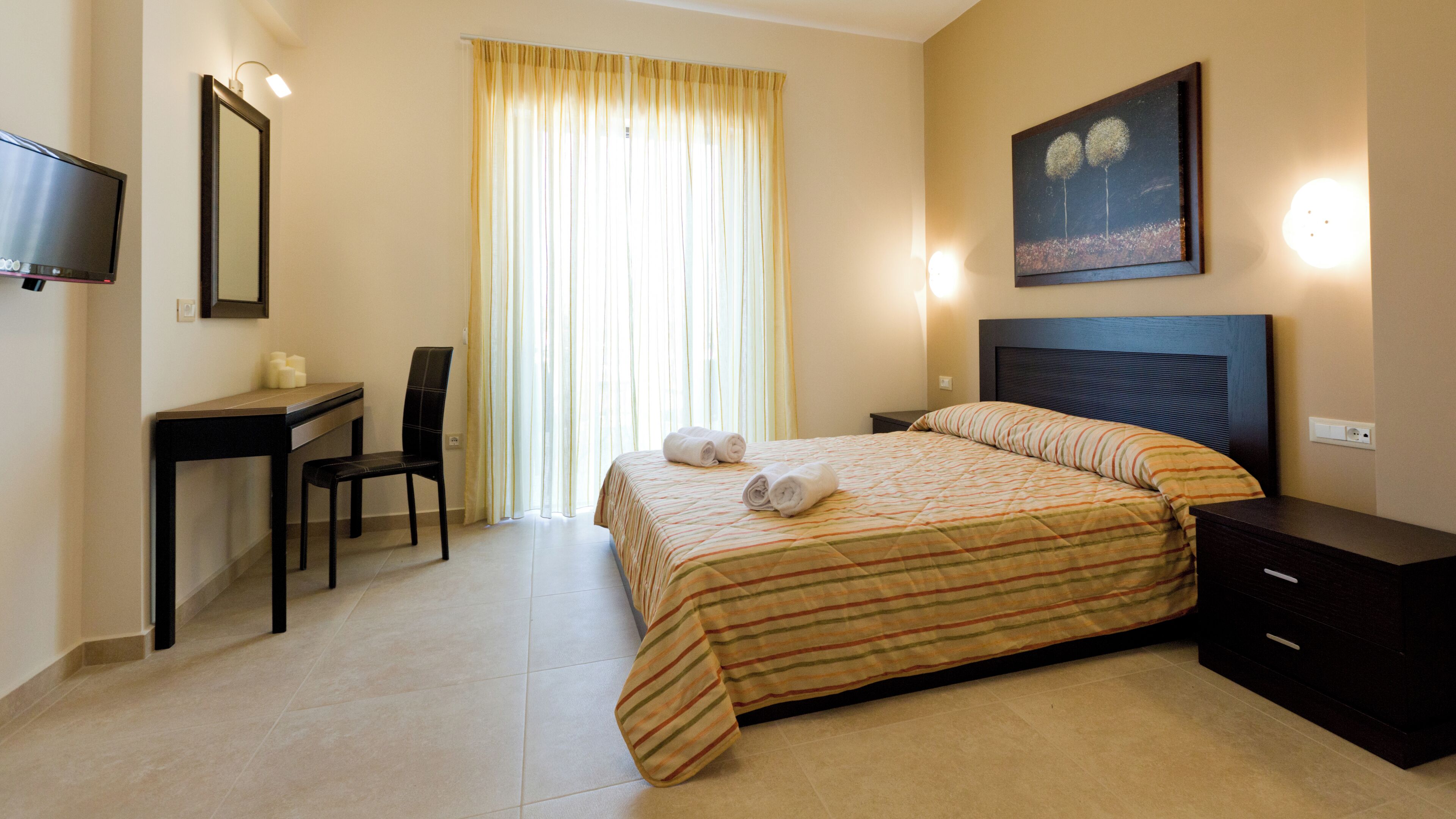 A hotel room with a bed, a desk, a chair, a TV, and a painting on the wall.