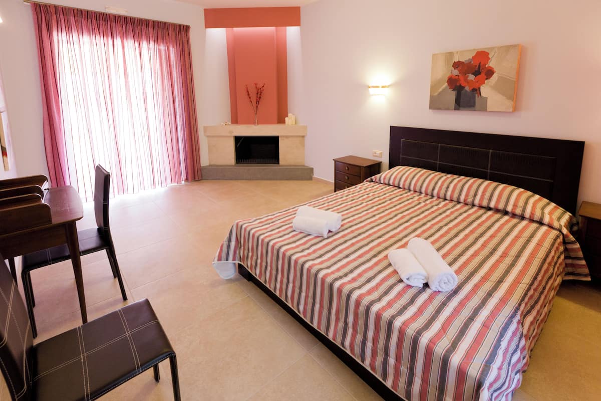 A hotel room with a bed, a chair, a desk, a fireplace, and a painting on the wall.