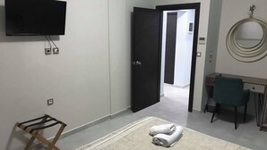 Habitación de hotel con televisión de pantalla plana montada en la pared, un escritorio con silla, un taburete de madera pequeño y una alfombra grande.