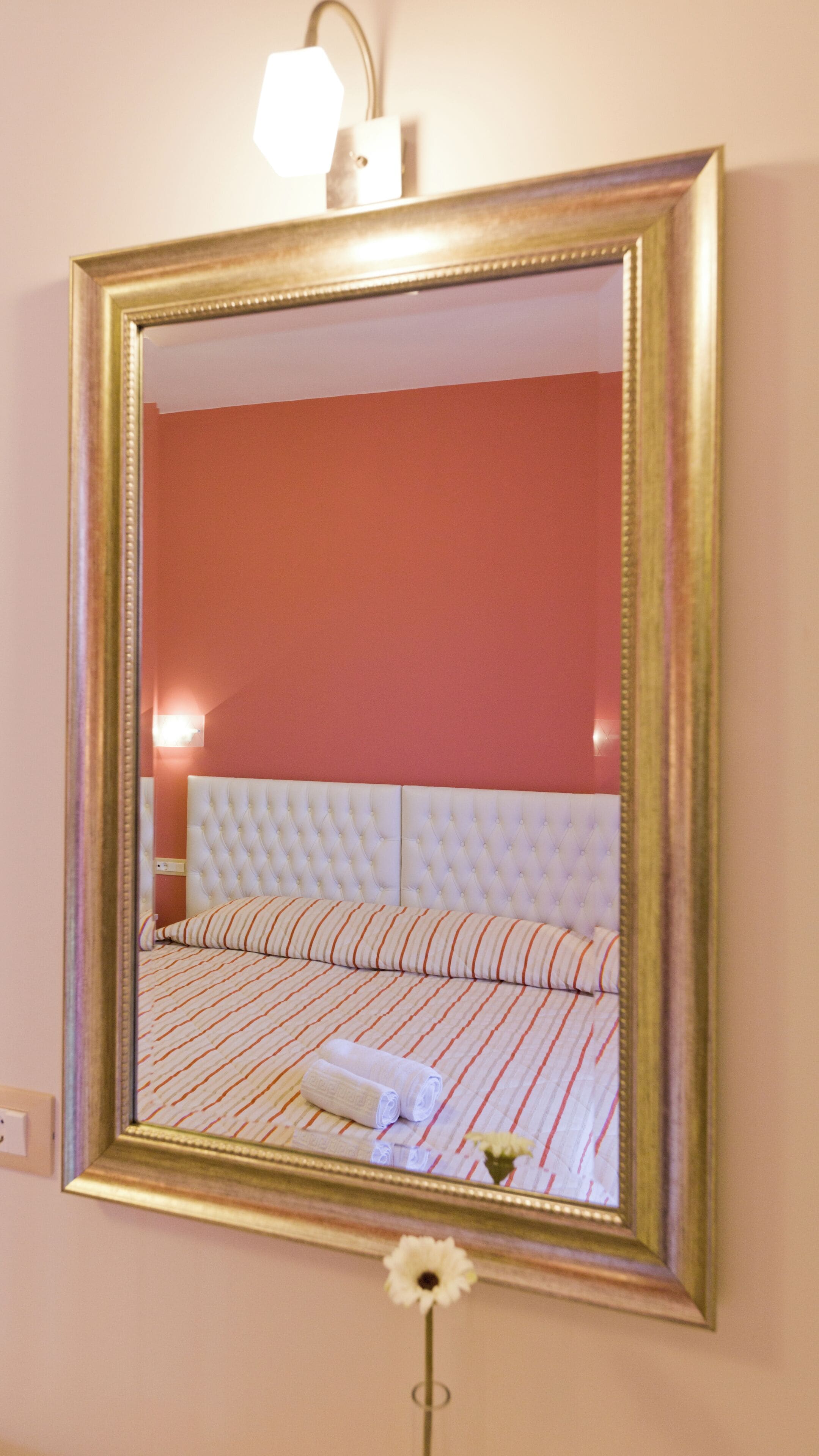 A mirror with a gold frame reflecting a room with a red wall, a white tufted headboard, striped bedding, and a flower on the floor.
