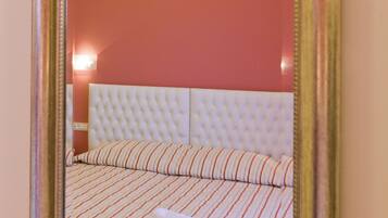 A mirror with a gold frame reflecting a room with a red wall, a white tufted headboard, striped bedding, and a flower on the floor.