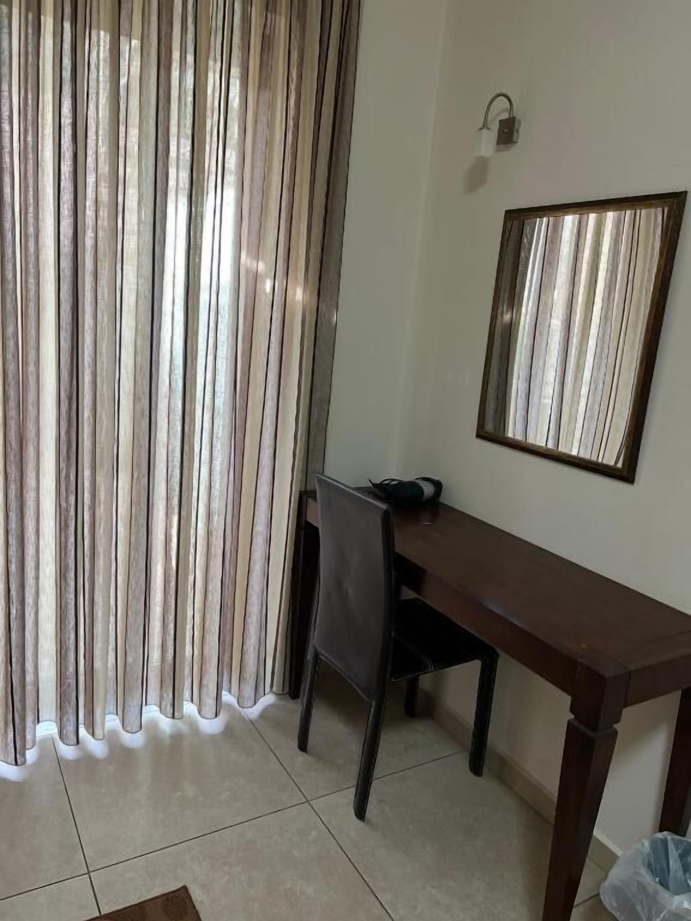 Une pièce comprenant un bureau en bois, une chaise, un luminaire fixé au mur et une fenêtre avec des stores verticaux.