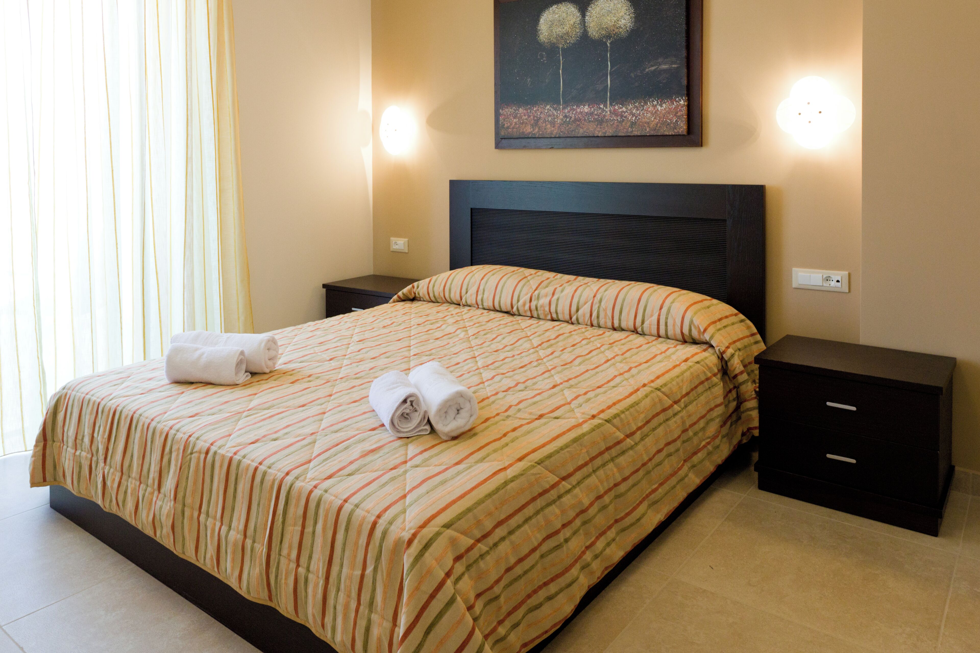 A neatly made bed with striped bedding and rolled towels, a dark headboard, and a nightstand with drawers.